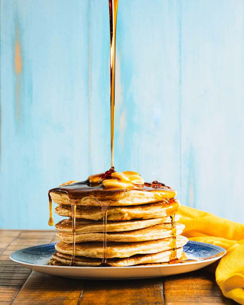 Big stack of butter-topped pancakes being drizzled with syrup